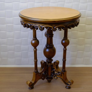 May include: A dark brown wooden side table with intricate carvings and a round top. The table has three legs with decorative ball finials.