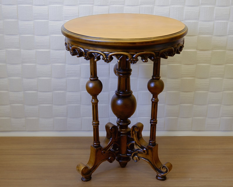 May include: A dark brown wooden side table with intricate carvings and a round top. The table has three legs with decorative ball finials.