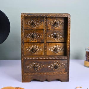 May include: A small, wooden jewelry box with seven drawers. The box is stained a warm brown color and features intricate carved geometric designs on the drawer fronts and frame. Each drawer has a small, round metal pull. The box is a decorative storage solution.