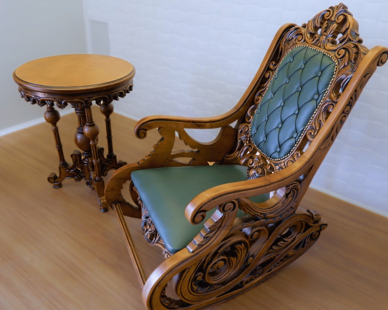 May include: A wooden rocking chair with intricate carvings and a green upholstered seat. A small wooden table with a round top sits next to the chair.