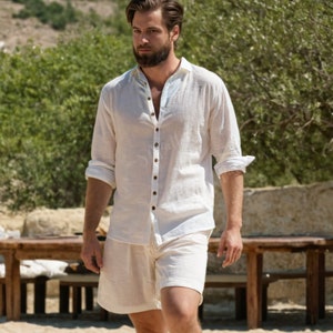 Puede incluir: Una camisa blanca de lino con cuello abotonado y mangas largas. La camisa se lleva sobre unos pantalones cortos de lino blancos.