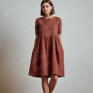 Puede incluir: Un vestido de lino de color óxido con cuello redondo y mangas cortas. El vestido tiene una cintura fruncida y dos bolsillos.