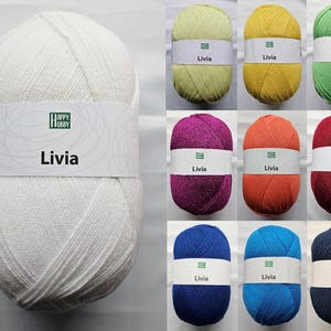 Könnte beinhalten: Eine Auswahl an Livia-Wollknäueln in verschiedenen Farben: Weiß, Gelb, Grün, Lila, Orange, Rot, Blau und Marineblau. Jedes Knäuel hat ein Etikett mit dem Happy Hobby-Logo und dem Namen "Livia". Ideal zum Stricken und Häkeln.