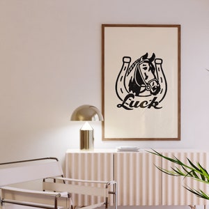 May include: A black and white print of a horse's head inside a horseshoe with the word "Luck" written underneath. The print is framed in a brown wood frame and hanging on a white wall.
