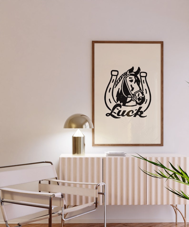 May include: A black and white print of a horse's head inside a horseshoe with the word "Luck" written underneath. The print is framed in a brown wood frame and hanging on a white wall.