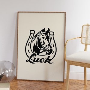 May include: Black and white illustration of a horse's head inside a horseshoe with the word "Luck" printed below. The illustration is on a beige background and framed with a light brown wood frame.