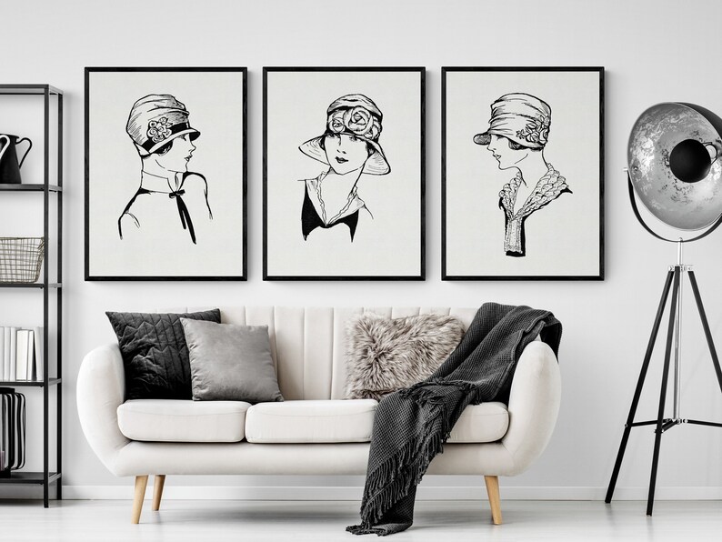 May include: Three black and white framed illustrations of women wearing hats. The illustrations are on a white wall above a white couch with black throw pillows and a black throw blanket.