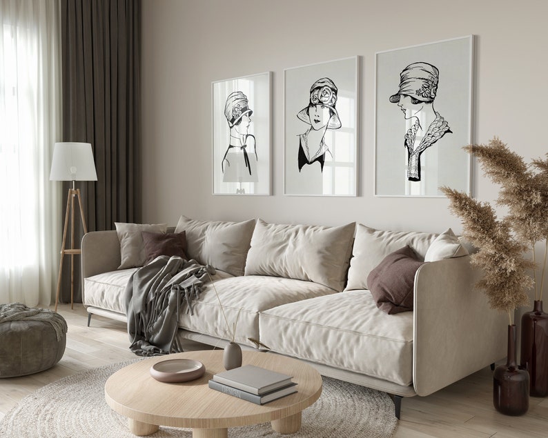 May include: Three black and white framed illustrations of women wearing hats, hanging on a light beige wall above a white couch with brown throw pillows and a blanket.
