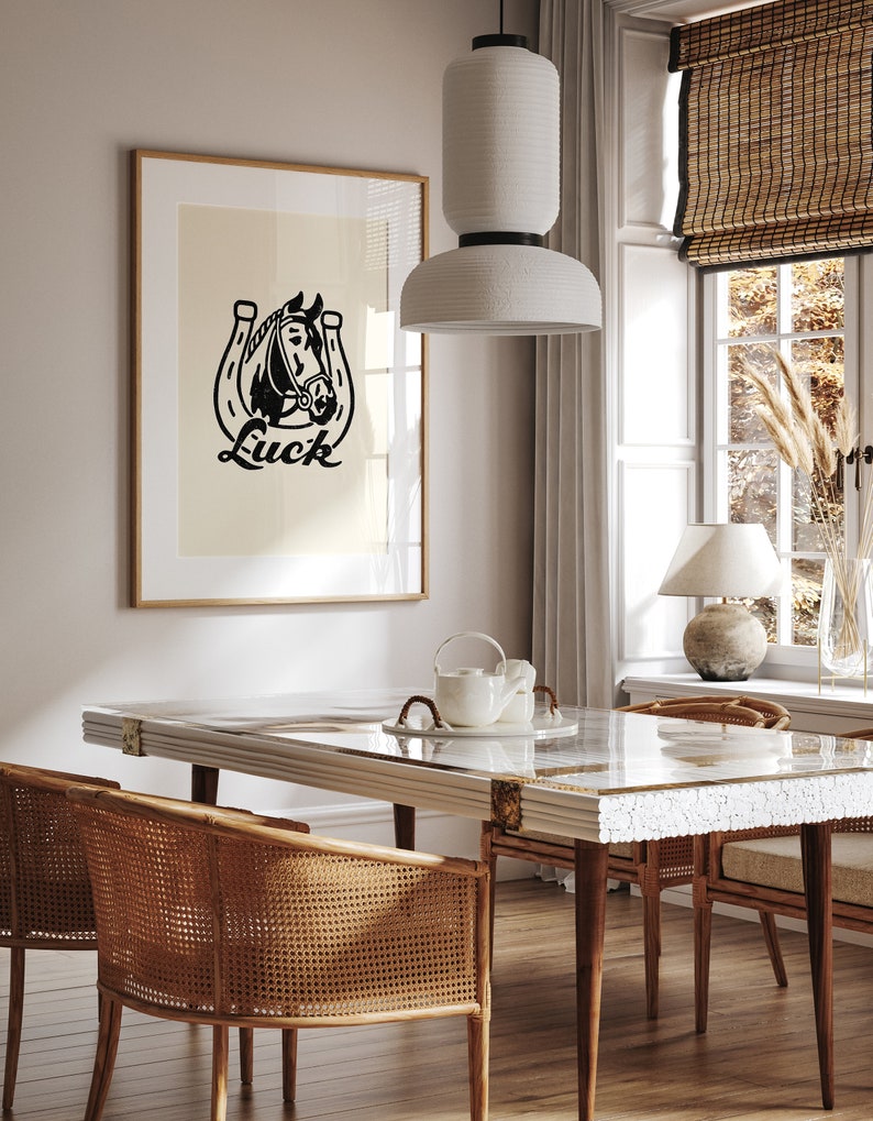 May include: A black and white illustration of a horse's head inside a horseshoe with the word "Luck" printed below. The illustration is framed and hanging on a wall in a dining room with a white table and wicker chairs.