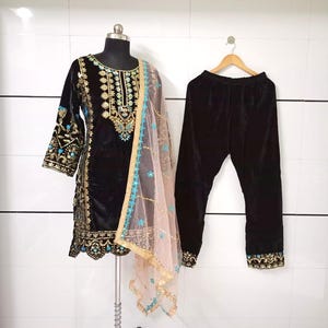 Può includere: Completo salwar kameez in velluto nero con ricami oro e turchese. Il set comprende una tunica a maniche lunghe, pantaloni abbinati e un dupatta rosa trasparente con finiture oro e turchese. La tunica è esposta su un manichino.