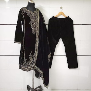 Puede incluir: Conjunto salwar kameez de terciopelo negro con intrincados bordados dorados. El conjunto incluye una túnica de manga larga, pantalones a juego y una bufanda drapeada. La túnica está en un maniquí, los pantalones en una percha de madera.