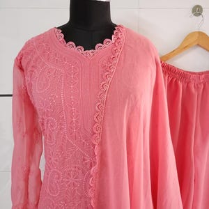 May include: A light coral-colored embroidered kurta suit displayed on a mannequin. The kurta features intricate floral and paisley patterns, long sleeves, and a scalloped neckline. A matching dupatta and pants are also visible.