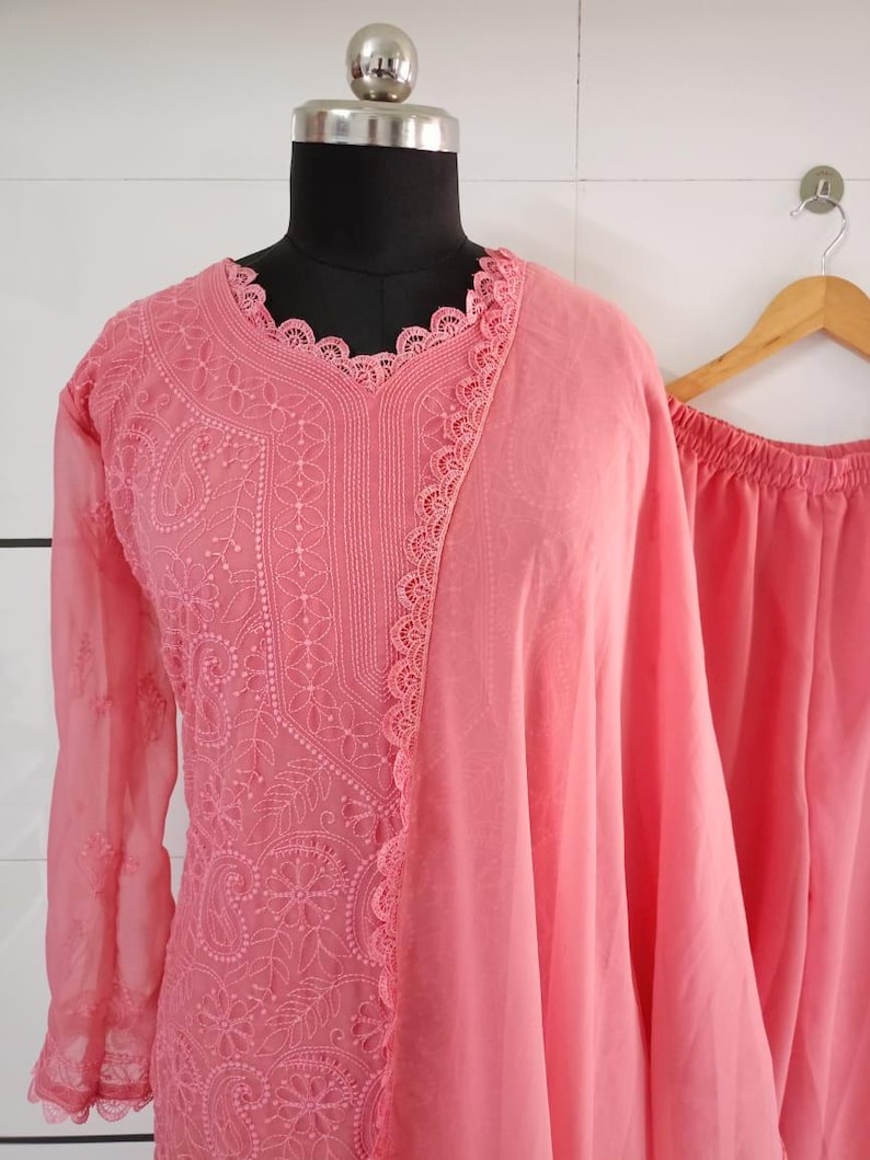 May include: A light coral-colored embroidered kurta suit displayed on a mannequin. The kurta features intricate floral and paisley patterns, long sleeves, and a scalloped neckline. A matching dupatta and pants are also visible.