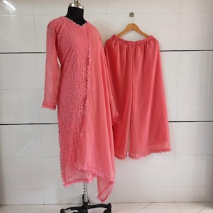 May include: A coral-colored, three-piece outfit displayed on a mannequin and hanger. The ensemble includes a long-sleeved tunic with intricate embroidery, a matching scarf, and wide-leg pants. The outfit is made of a lightweight fabric.