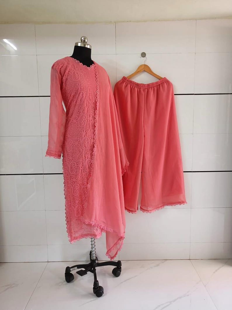 May include: A coral-colored, three-piece outfit displayed on a mannequin and hanger. The ensemble includes a long-sleeved tunic with intricate embroidery, a matching scarf, and wide-leg pants. The outfit is made of a lightweight fabric.