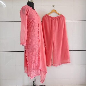 May include: A coral-colored, two-piece outfit. The top is a long-sleeved, open-front tunic with intricate lace detailing. Matching wide-leg pants hang on a wooden hanger. The outfit is displayed on a mannequin and a hanger against a white tiled wall.
