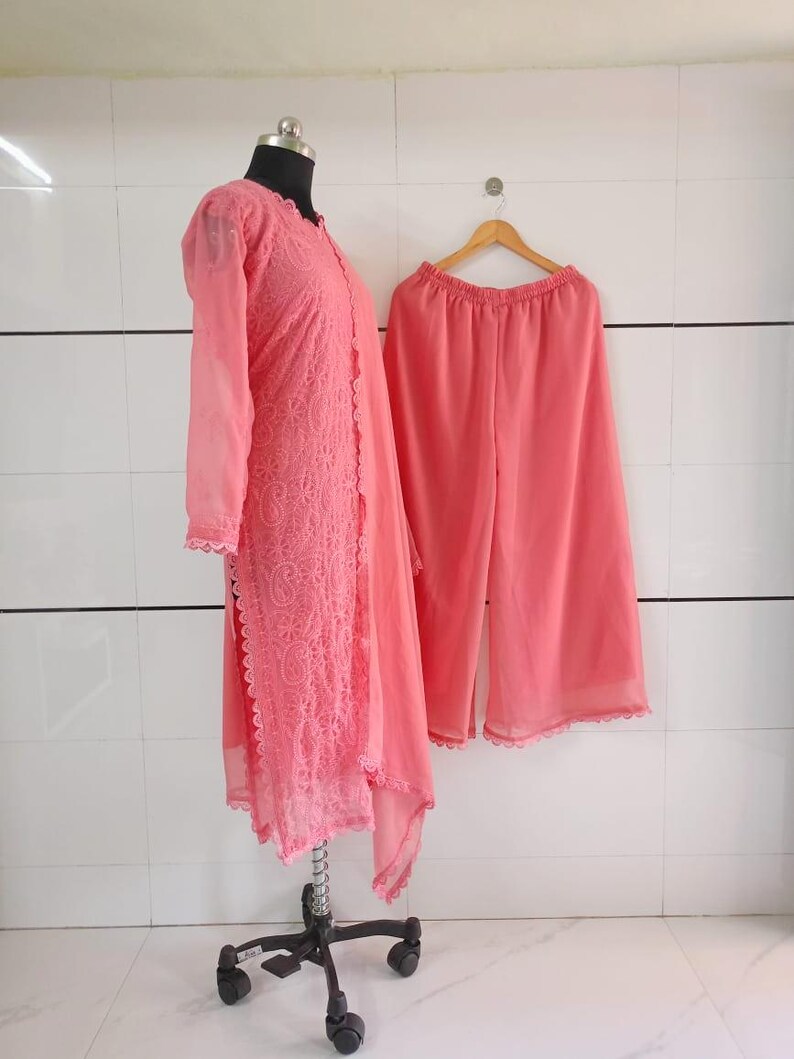 May include: A coral-colored, two-piece outfit. The top is a long-sleeved, open-front tunic with intricate lace detailing. Matching wide-leg pants hang on a wooden hanger. The outfit is displayed on a mannequin and a hanger against a white tiled wall.