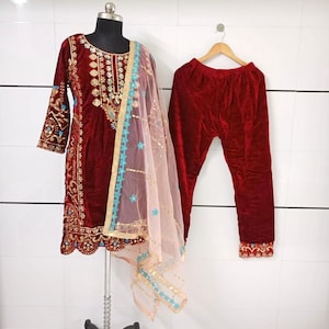 May include: A burgundy velvet salwar kameez set. The set includes a long-sleeved tunic with intricate gold embroidery and a matching pair of pants. A sheer, peach-colored scarf with blue and gold trim completes the ensemble. The outfit is displayed on a mannequin.