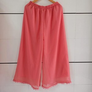 May include: Coral-colored palazzo pants hanging on a wooden hanger. The wide-leg pants have an elastic waistband and a scalloped lace trim at the hem. The pants are made of a lightweight, sheer fabric.