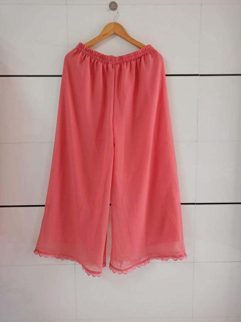 May include: Coral-colored palazzo pants hanging on a wooden hanger. The wide-leg pants have an elastic waistband and a scalloped lace trim at the hem. The pants are made of a lightweight, sheer fabric.