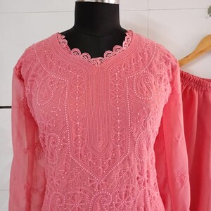 May include: A coral-colored, embroidered tunic and matching pants set displayed on a mannequin. The tunic features intricate floral and paisley patterns, long sleeves, and a scalloped neckline. The fabric appears lightweight and semi-sheer, with a delicate, detailed design.