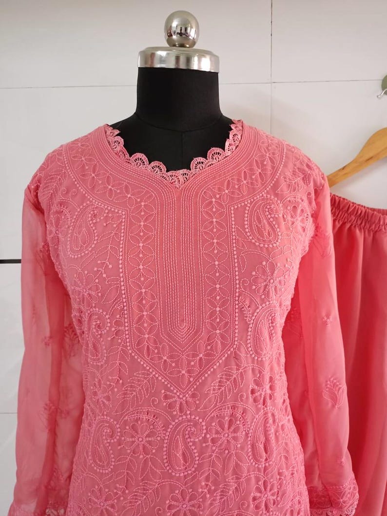 May include: A coral-colored, embroidered tunic and matching pants set displayed on a mannequin. The tunic features intricate floral and paisley patterns, long sleeves, and a scalloped neckline. The fabric appears lightweight and semi-sheer, with a delicate, detailed design.