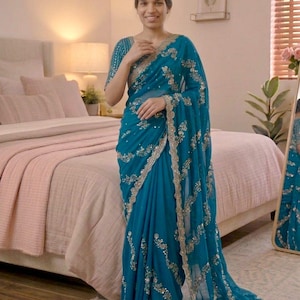 May include: A teal saree featuring intricate silver embroidery and sequin detailing along the borders and throughout the fabric. The garment includes a matching short-sleeved blouse with coordinated silver embellishments on the sleeves and neckline.