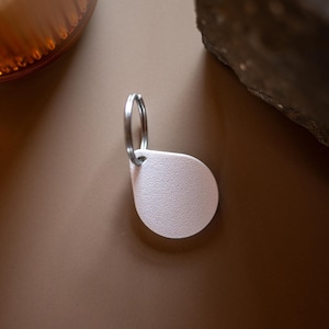 Könnte beinhalten: Ein hellrosa, tropfenförmiger Schlüsselanhänger mit strukturierter Oberfläche. Der Anhänger ist an einem silbernen Metall-Schlüsselring befestigt. Der Hintergrund ist einfarbiges, gedämpftes Braun.