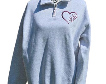 May include: A light gray sweatshirt with a quarter-zip collar. The sweatshirt features a burgundy heart-shaped embroidered design with the name "Lili" inside. The long-sleeved top is displayed on a mannequin.