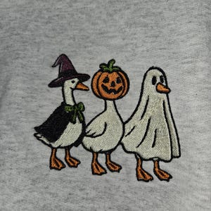 May include: Embroidered design featuring three geese dressed for Halloween. One goose wears a witch hat and cape, another a pumpkin head, and the last is in a ghost costume. The design is on a light gray background.