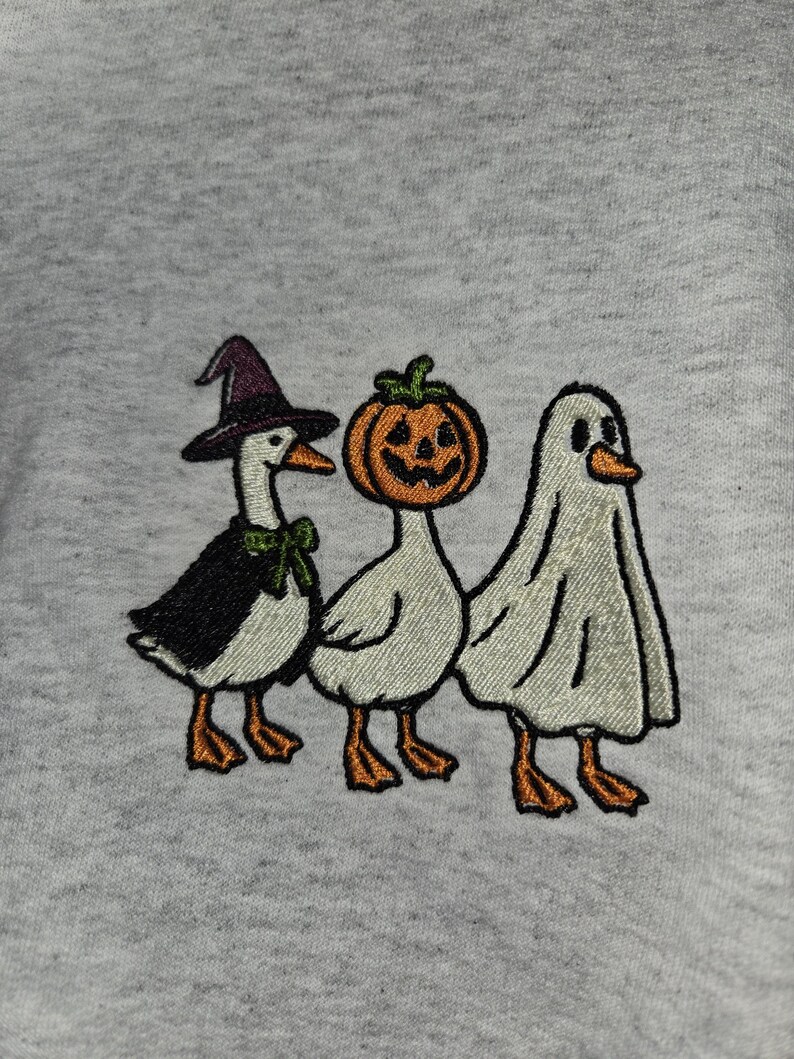 May include: Embroidered design featuring three geese dressed for Halloween. One goose wears a witch hat and cape, another a pumpkin head, and the last is in a ghost costume. The design is on a light gray background.