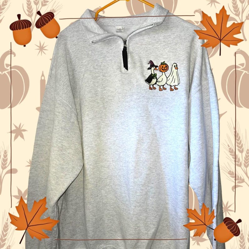 May include: A light gray sweatshirt with a quarter-zip neckline and a small embroidered design of three cartoon geese in Halloween costumes. The geese are dressed as a witch, a jack-o'-lantern, and a ghost. The sweatshirt is hanging on a gold hanger.