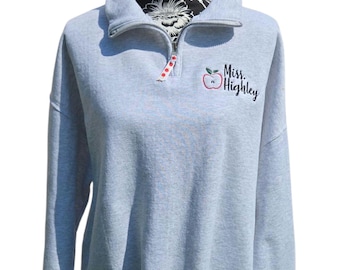 May include: A light gray pullover sweatshirt with a quarter-zip neckline. The sweatshirt features a red apple graphic and the text "Miss. Highley" in black script. The zipper pull has a red and white polka dot design.