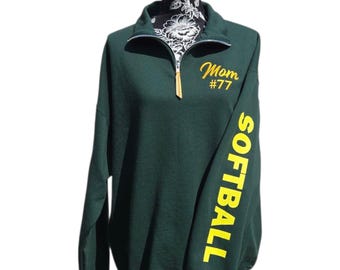May include: Dark green sweatshirt with a quarter-zip collar. The sleeve features the word "SOFTBALL" in yellow block letters. The chest has the text "Mom #77" in yellow script. The sweatshirt is displayed on a black mannequin.