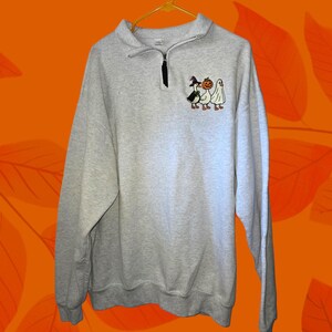 May include: A light gray sweatshirt with a quarter-zip neckline and a small embroidered design of three Halloween characters: a witch goose, a pumpkin, and a ghost. The sweatshirt is hanging on a yellow hanger.