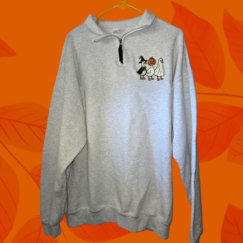 May include: A light gray sweatshirt with a quarter-zip neckline and a small embroidered design of three Halloween characters: a witch goose, a pumpkin, and a ghost. The sweatshirt is hanging on a yellow hanger.