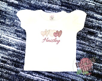 May include: White t-shirt with a red embroidered design of four patchwork hearts and the name "Haisley" below.