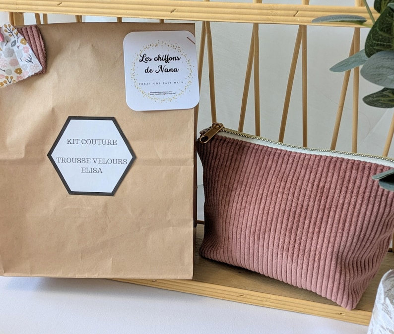 May include: A brown paper bag with a black and white label that reads "KIT COUTURE TROUSSE VELOURS ELISA" is next to a pink corduroy pouch with a gold zipper. A small white card with the text "Les chiffons de Nana" is also visible.