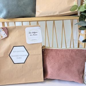 May include: A display of corduroy pouches and paper bags. The pouches are in shades of dusty rose and sage green, with a vertical ribbed texture. The paper bags have labels with text. The items are arranged on a light-colored shelf.
