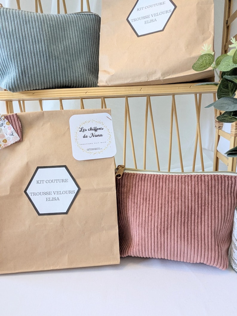 May include: A display of corduroy pouches and paper bags. The pouches are in shades of dusty rose and sage green, with a vertical ribbed texture. The paper bags have labels with text. The items are arranged on a light-colored shelf.