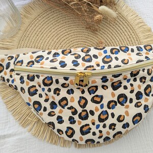 Puede incluir: Una riñonera blanca con un diseño de estampado de leopardo en tonos marrones, negros y azules. La riñonera tiene una cremallera dorada.