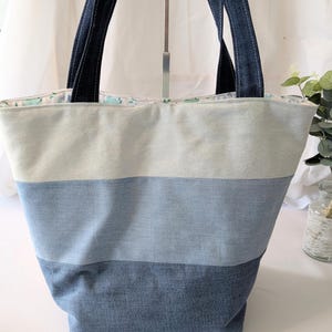 Peut inclure: Un sac fourre-tout en denim avec trois rayures horizontales de différentes nuances de bleu. Le sac a des poignées en denim bleu foncé et une doublure à motifs floraux. Le sac est conçu pour transporter des objets.