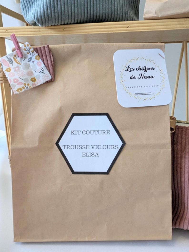 May include: A brown paper bag labeled "KIT COUTURE TROUSSE VELOURS ELISA" with a black and white hexagon-shaped label. A small fabric swatch with floral patterns and a pink corduroy swatch are clipped to the bag. A white tag with gold text reads "Les chiffons de Nana".