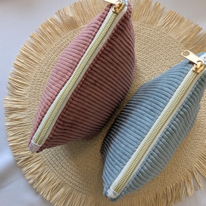 May include: Two corduroy pouches, one dusty rose and one light blue, with gold-toned zippers. The pouches are on a round, woven placemat with a fringed edge. The pouches are a soft, textured material.