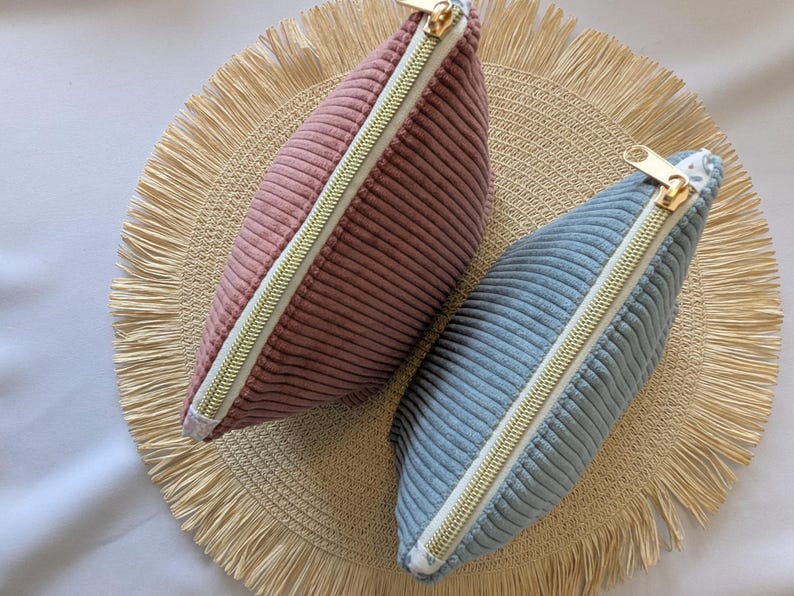May include: Two corduroy pouches, one dusty rose and one light blue, with gold-toned zippers. The pouches are on a round, woven placemat with a fringed edge. The pouches are a soft, textured material.