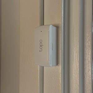 Puede incluir: Un dispositivo doméstico inteligente Tapo blanco montado en una puerta beige. El dispositivo tiene la marca "tapo" impresa. El dispositivo es rectangular con una parte delantera ligeramente curvada y una pequeña luz indicadora.