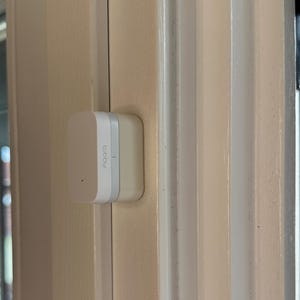 Può includere: Un dispositivo Aqara per la casa intelligente, bianco e grigio chiaro, montato su un telaio di porta beige chiaro. Il dispositivo ha una forma rettangolare con angoli arrotondati e il nome del marchio stampato.