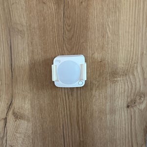 Può includere: Un sensore di movimento Ring bianco con una luce circolare, montato su una superficie in legno. Il dispositivo è rettangolare con angoli arrotondati e ha il marchio "ring" stampato. La superficie in legno presenta un motivo a venature naturali.