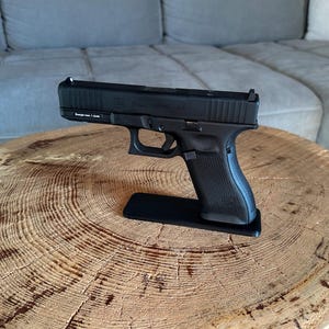 May include: A black semi-automatic pistol with a textured grip and a matte finish, mounted on a small black rectangular stand. The slide features the text Glock 17 AUSTRIA 9x19 and a small label on the frame reading Energie max. 1 Joule.