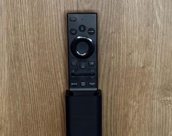 Może przedstawiać: Czarny pilot do telewizora z okrągłym panelem nawigacyjnym i przyciskami do Netflix, Prime Video i TV Plus. Pilot jest zamontowany na czarnym uchwycie ściennym.
